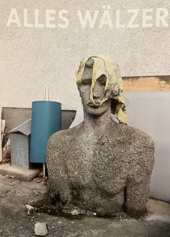 lebensgroße männliche Stein-bzw Betonbüste, die, wie es scheint, aus dem Asphalt wächst; über dem Kopf ein schmutziges Unterleiberl,dahinter eine türkise Kunststoffrolle ua. abgelegte Gegenstände