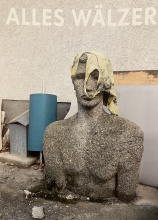 lebensgroße männliche Stein-bzw Betonbüste, die, wie es scheint, aus dem Asphalt wächst; über dem Kopf ein schmutziges Unterleiberl,dahinter eine türkise Kunststoffrolle ua. abgelegte Gegenstände